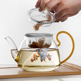 Glass teapot with a decorative handle, filled with tea, on a wooden surface.