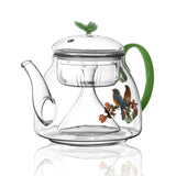 Clear glass teapot with green handle and bird design on a white background