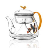 Clear glass teapot with orange handle and bird design on a white background