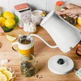 Person pouring tea from a white kettle into a glass jar on a wooden table with lemons and sugar in the background.