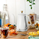White electric kettle on a wooden table with a glass mug of tea, oranges, and a bowl of lemons.